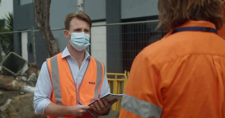 A man with a vest mask pen and notebook standing and talking to another man