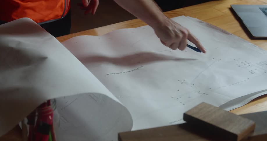 A person in a safety vest marks a blueprint lying on a desk with a pen