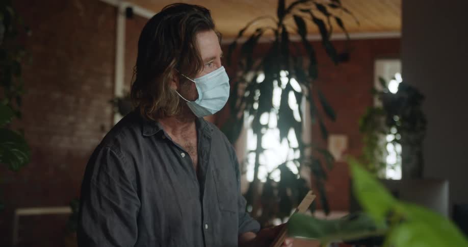 A man with a mask talking in a room with plants and windows in the background