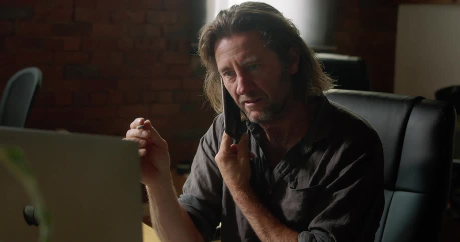 A man speaks on the phone while sitting at a desk in front of a computer and paper