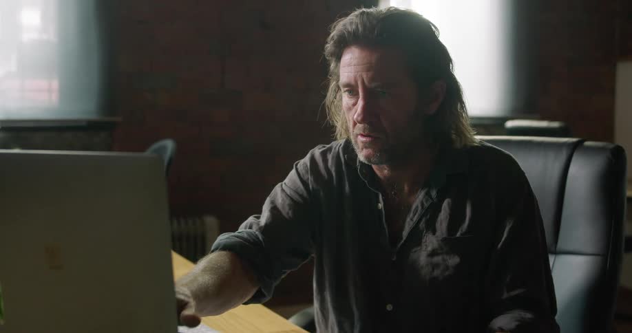 A male sitting at a desk working on a computer flipping through a book
