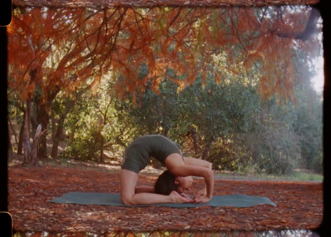 A woman is kneeling on a mat under a tree in a leaf covered yard as she raises her arms and bends backwards and then holds her ankles with her hands