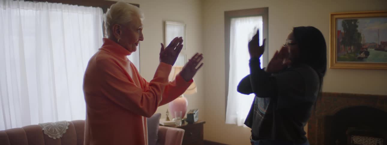 An elderly woman at home mimics motions that a young black woman has her do