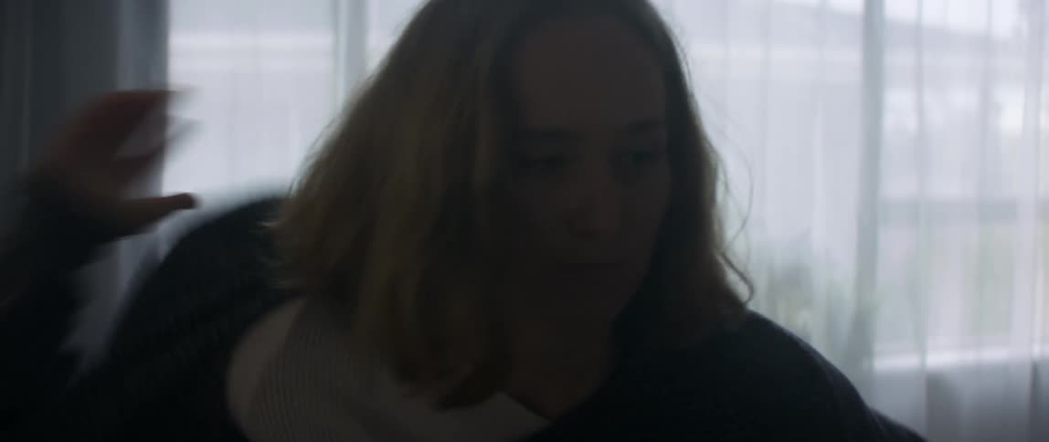 Blonde woman puts on gray sweater sitting on bed in bedroom