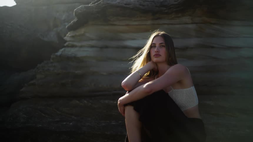 Woman with brown hair white top and black skirt sits near big rock on the coast
