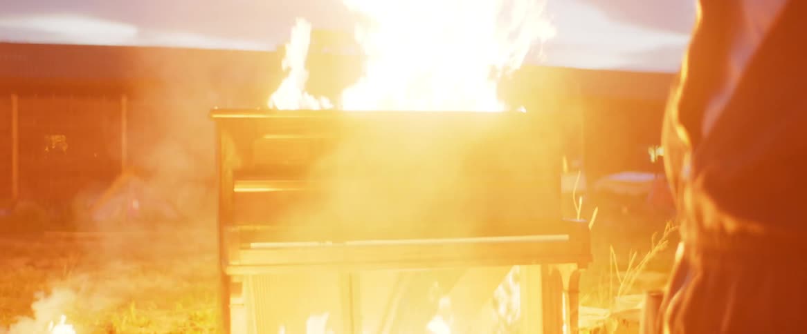 A teenage boy is standing outside of a nearby building holding a matchbox and lighting things on fire as he strikes a match and throws it on the ground as an old piano bursts into flames