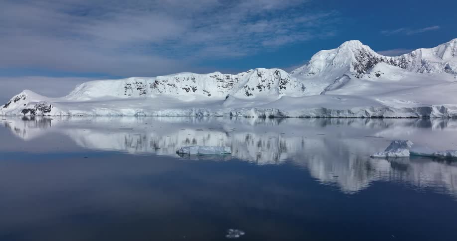 Sea ice floats on water reflecting snowy mountains near coast under a cloudy day
