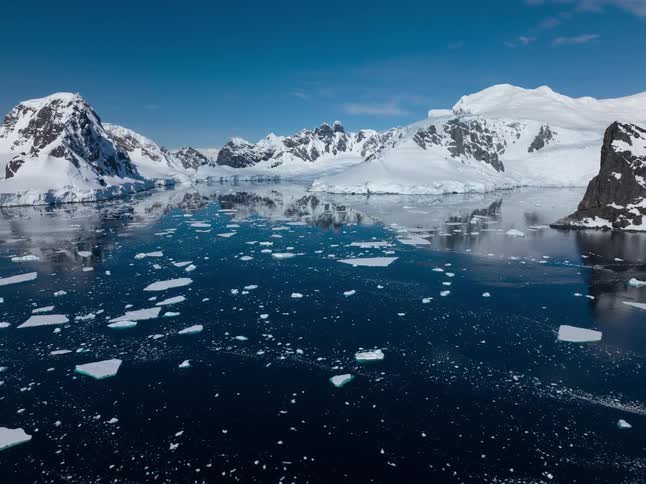 Snow covered rocky shore is reflected on the smooth blue water surface with icebergs moving in hyperlapse