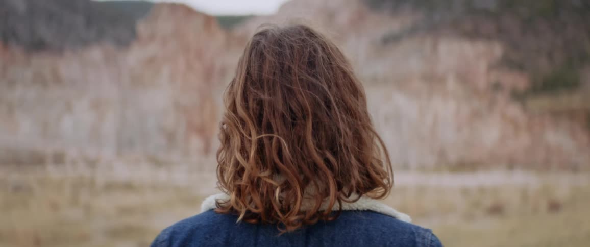Rear view of woman with brown wavy hair and denim jacket watching the large rocks