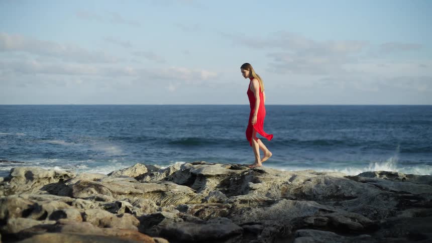 Woman with brown hair and red dress walks barefoot on rocky shore near the ocean