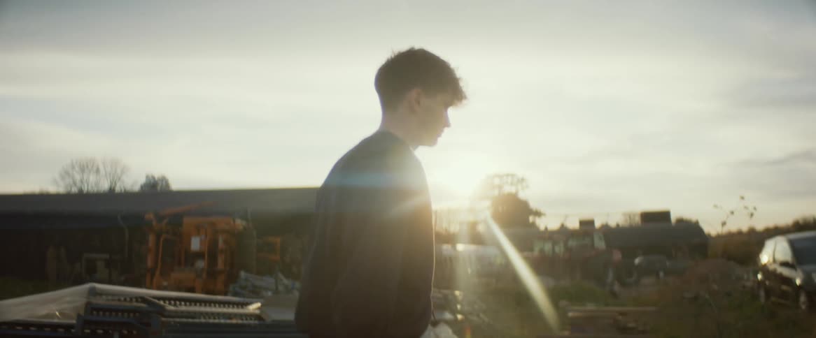 The sun shines brightly over a building in a rural area as a teenage boy carrying a stick is walking toward a vehicle