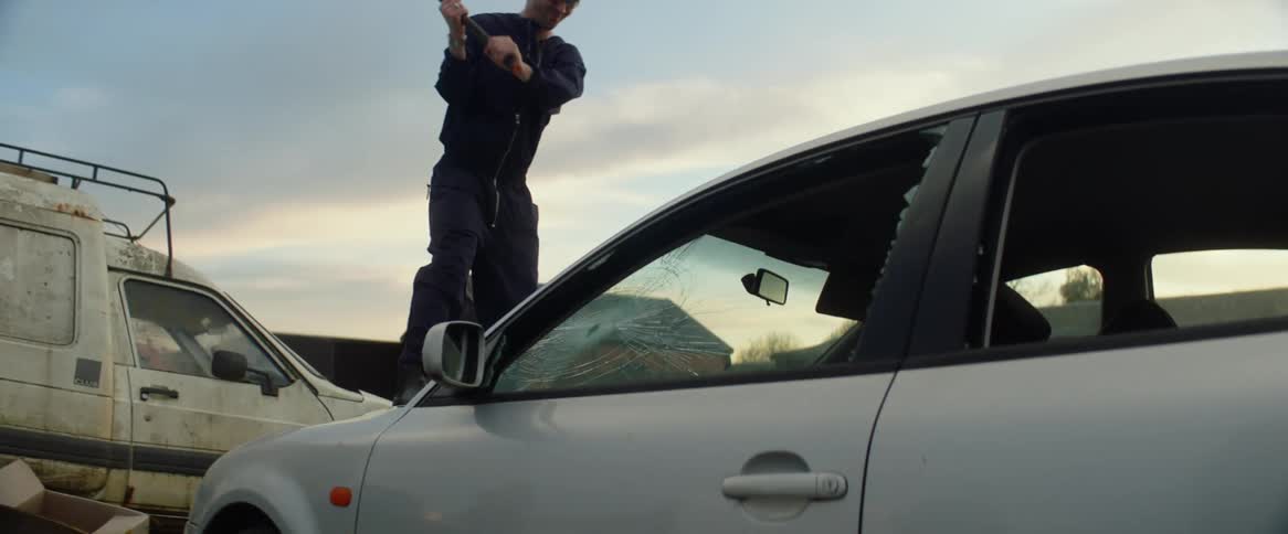 A man with a hammer standing on a car and smashing the windshield with thehammer