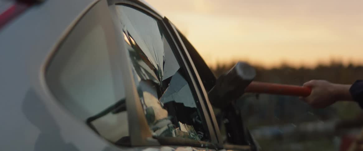 Person smashes a car window with a sledge hammer at dusk