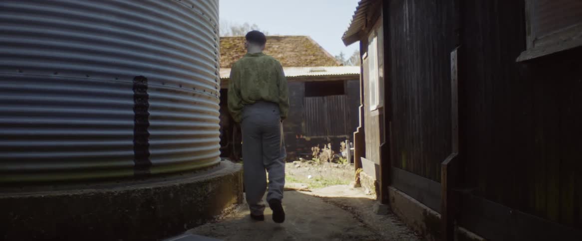 A teenage boy is walking with his hands in his pockets near building and he stands leaning against a silo