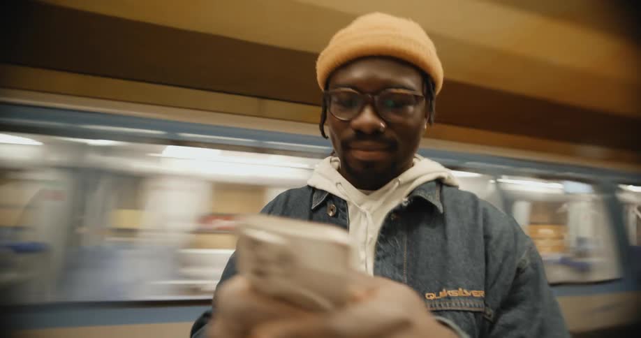 Man smiles while viewing smartphone in terminal as a subway train passes in the background
