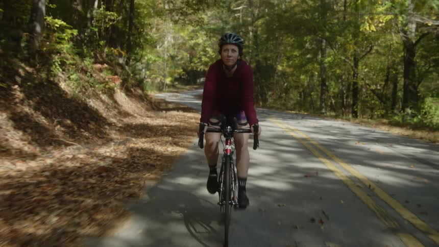 A woman rides her bicycle down a winding road past a forest