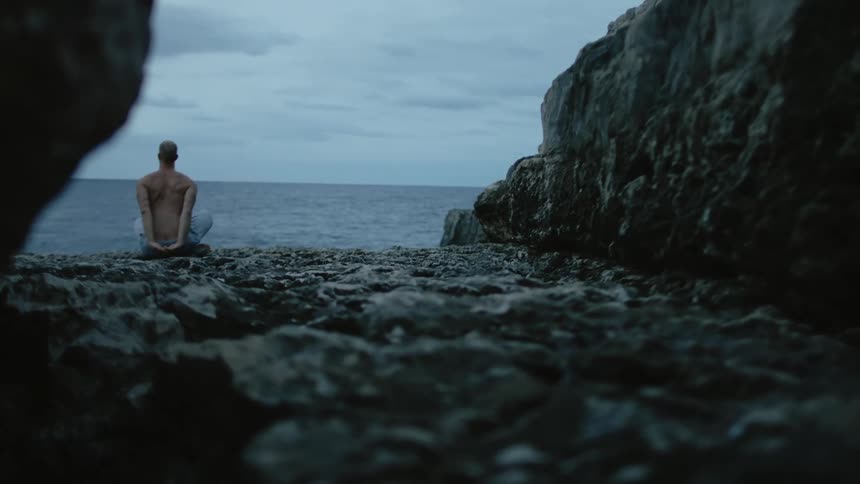 A man is sitting on rocks along the coast with his hands behind his bare back as he moves his arms