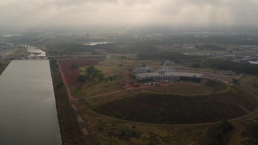 Automobiles are parked at a facility in a round mound next to a lake as traffic moves on the highway under a foggy cloudy sky