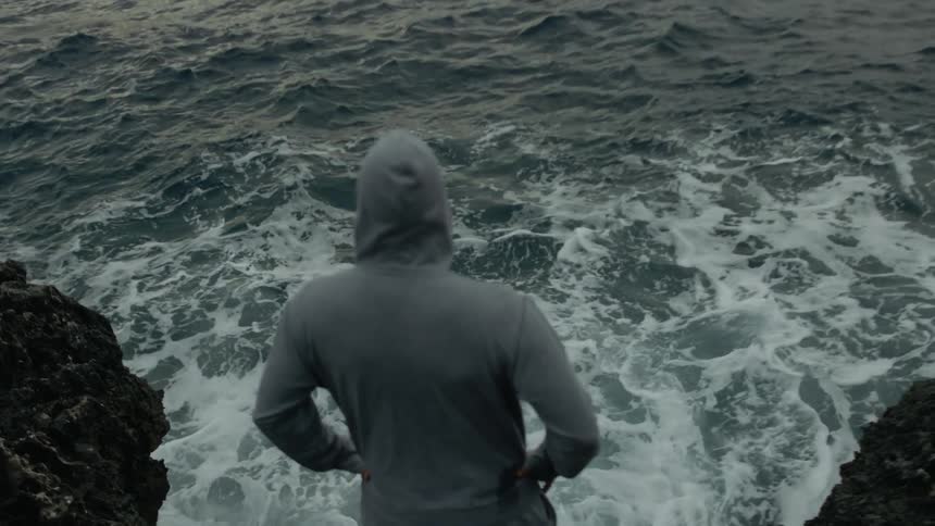 A man wearing a hoodie is standing on rocks along the coast watching the water was it waves and moves