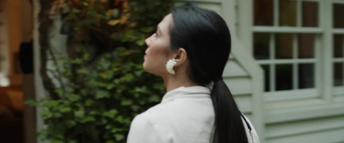 A woman with long black hair tied back in a pony tail is looking around as she walks to a house and goes inside the open front door