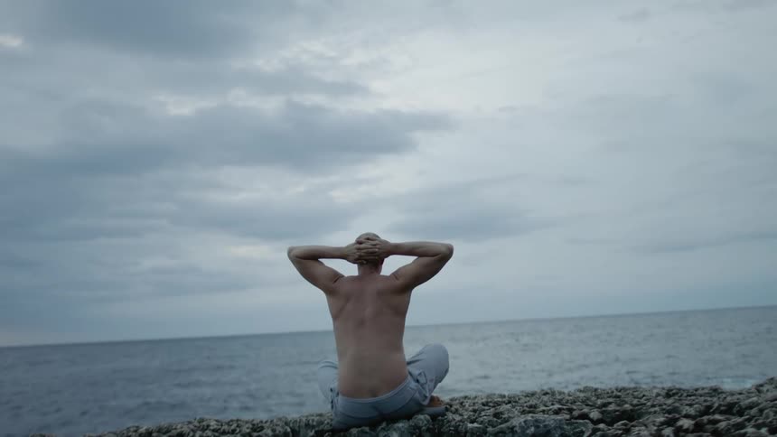 A man is sitting on rocks along the coast with his hands clasped behind his head as he watches the waves roll into shore