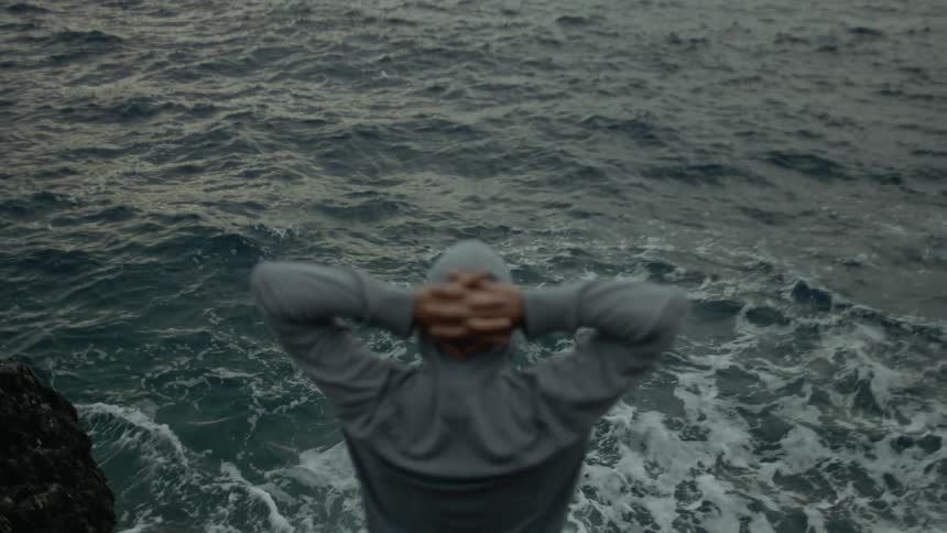 A man stands above crashing waves in the ocean and raises his arms above his head and clasps his hands behind his neck