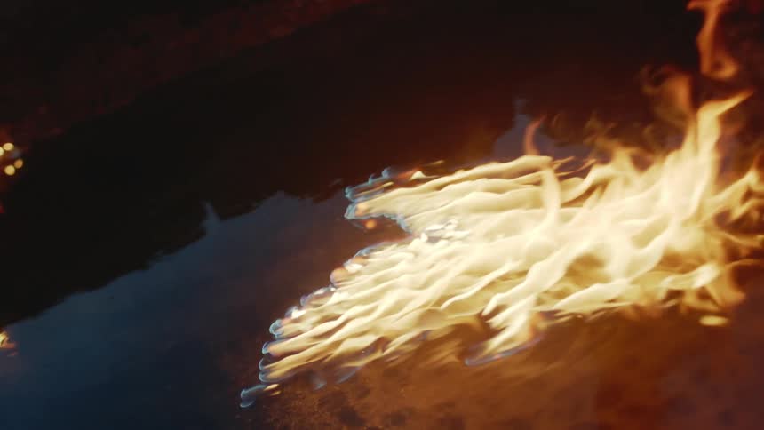 Tall flames are moving across the surface on a body of water with the lands edge close by