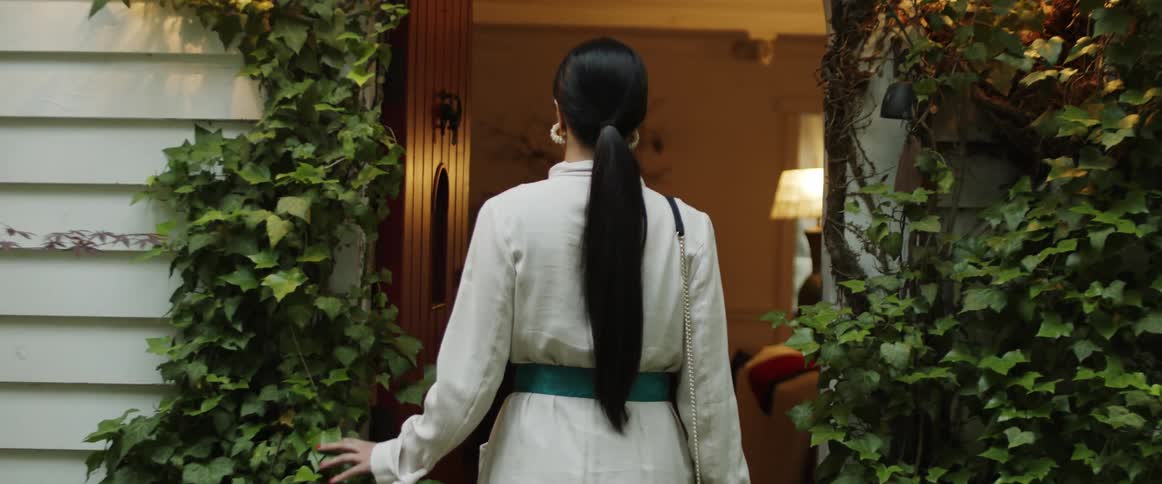 A woman with long dark hair walking into a house with green vines lining the doorway