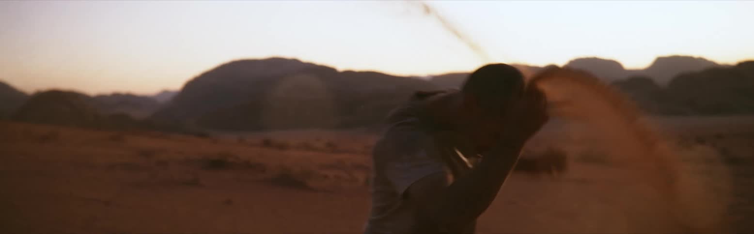 Man scrambles on the ground tossing red sand onto himself with dirt hills in the distance