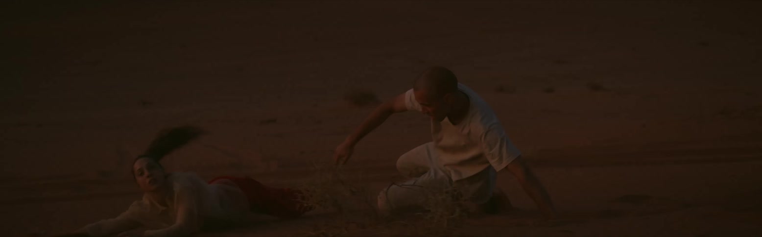 A man and a woman are on the ground in the desert and he is rolling her across the sand