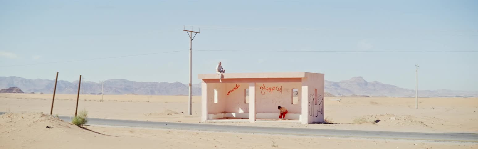 One person sits on roof and another on bench of graffiti covered bus stop near road in desert