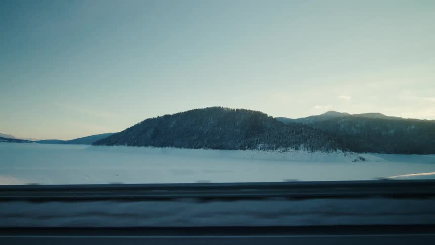 Pov from car passenger side driving on highway looking at snowy landscape passing by and mountains in the background