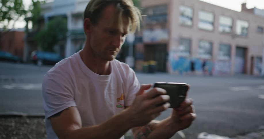 A man is kneeling down on the side of a downtown street taking a picture with his phone