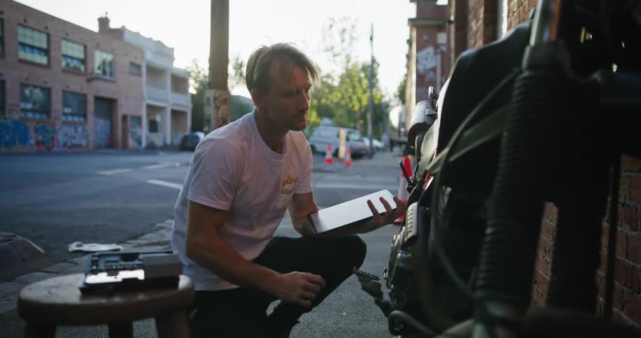 Man consults tablet while working on motorcycle across from road construction