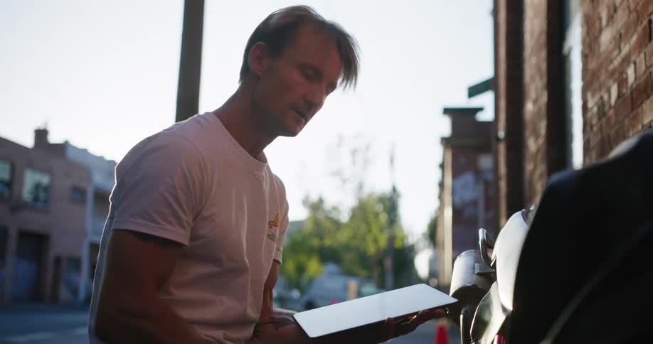 A man swipes through a tablet while looking at a motorcycle he seems to be working on