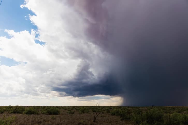 Dark storm clouds move in rotation over a field as rain pours down from the clouds and lightning flashes