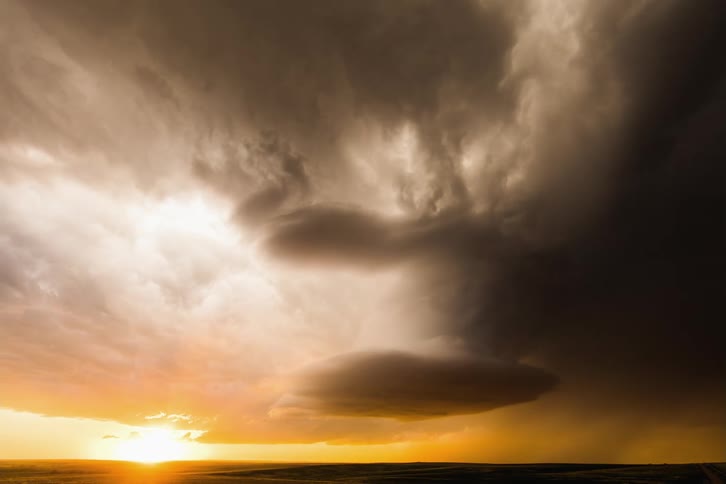 Dark storm clouds are moving across an orange sky as lightning flashes and the sun is on the horizon