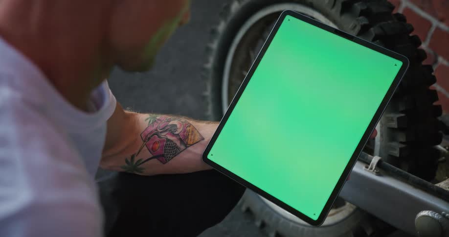 Man near motorcycle parked close to brick wall views tablet