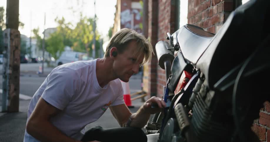 Blonde man with white shirt and black pants fixes motorcycle in the street