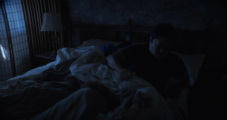 A couple are lying in bed asleep as the man wakes up and sits on the side of the bed