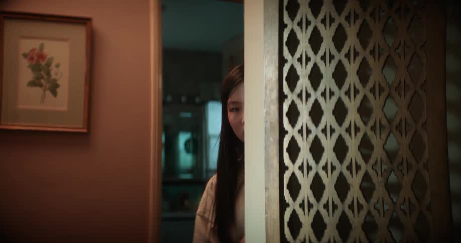 A teenage girl is standing in a hallway carefully peeking around the corner