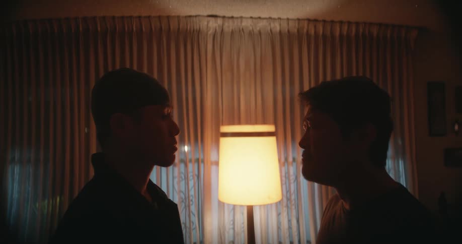 Older man stands face to face with younger man in dimly lit home