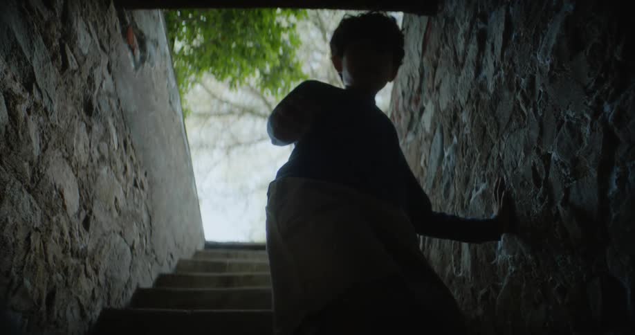 Child walks down stairs enclosed between stone walls and is silhouetted against opening while reaching down