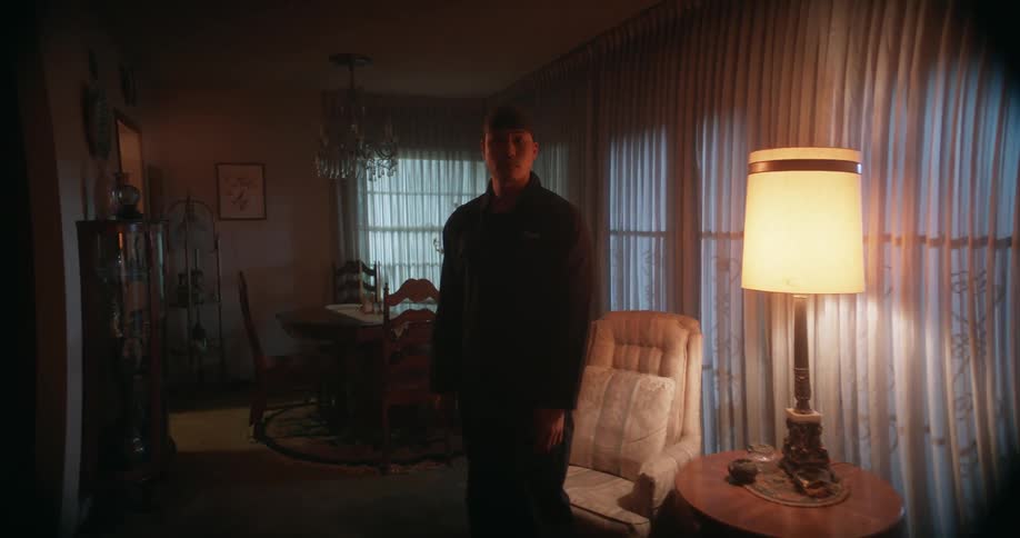 A man is standing silently in the living room in front of a chair next to a table lamp that is on