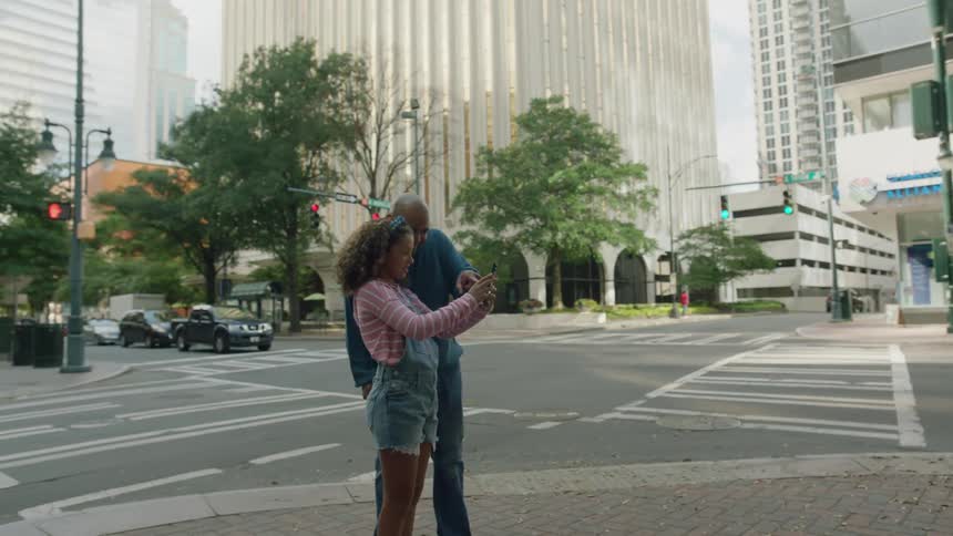 A young girl is standing on a downtown street with her father as she holds up her phone and he points to the screen