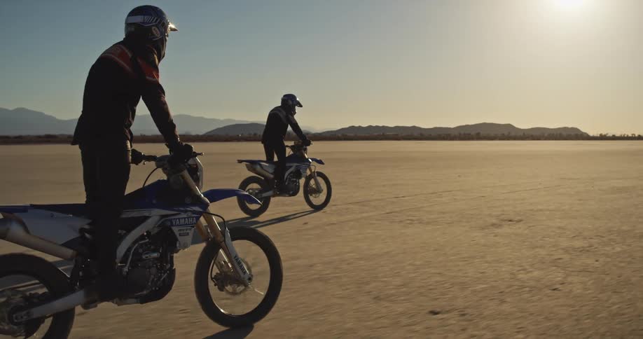 Two people riding dirt bikes through the sandy desert with mountains in the distance