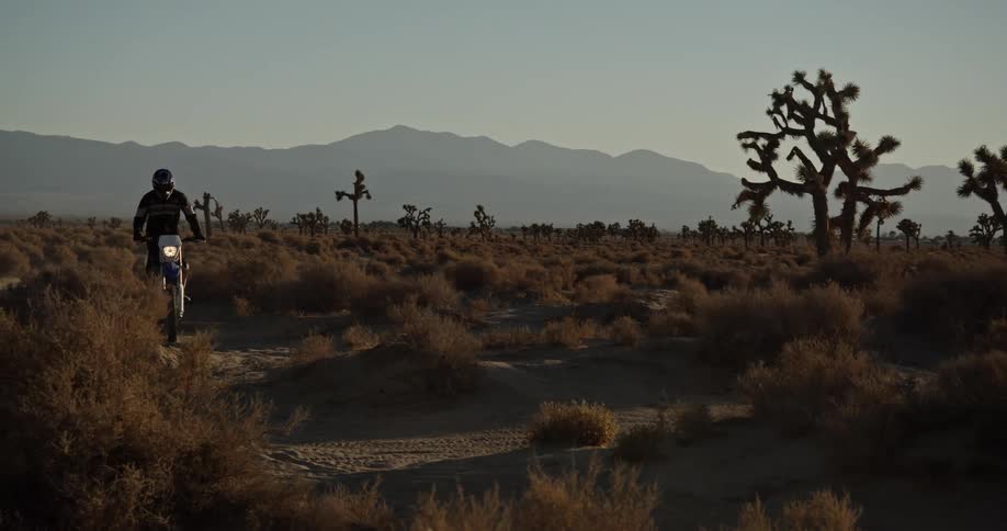 Motorcyclist wearing helmet and jacket rides bike across arid scrub land with misty mountains in the background