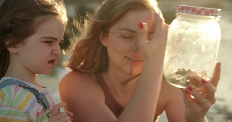 A woman is holding a glass jar and showing her young daughter as she points to something inside as waves splash over rocks along the shoreline