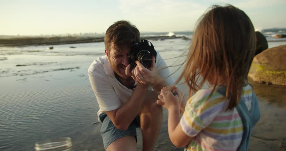A father takes a picture of his daughter holding something on the beach while the mom holds a jar
