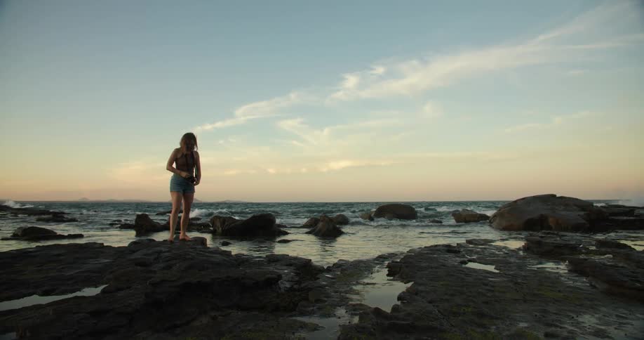 Woman takes picture while standing on shore against a red tinted horizon